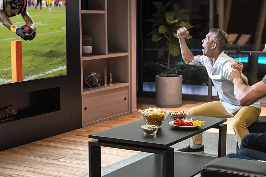 Men cheering while watching football on large TV in living room with food on coffee table